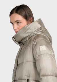 Ljusgrå vadderad jacka med hög krage, med en glansig yta och en logotypmärke på axeln. Dragkedja framtill och quiltad design.