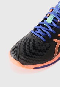 Chaussure de sport noire avec un dessus texturé, semelle orange et lacets bleus. Présente un accent vert sur la languette et des bandes sur le côté.