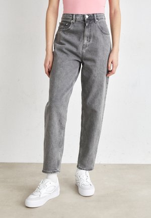 Person, die graue Jeans mit hoher Taille, weiße Socken und weiße Sneakers trägt, steht auf einem schlichten hellen Fußboden vor einer weißen Wand.