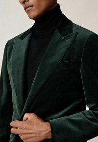 Roheline sametblazer, millel on šaalikaelus, ühekohaline nööp kinnitamiseks ja tasku detailid. Kandmine musta kõrge kaelusega särk üle, et luua kihiline välimus.