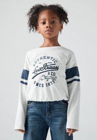 Nuori tyttö, jolla on kihara hius, on pukeutunut valkoiseen pitkähihaiseen paitaan, jossa on tummansinisiä raitoja ja tekstiä, sekä sinisiin farkkushortseihin. Hän seisoo yksinkertaista taustaa vasten.