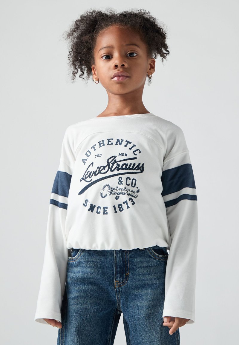 Nuori tyttö, jolla on kihara hius, on pukeutunut valkoiseen pitkähihaiseen paitaan, jossa on tummansinisiä raitoja ja tekstiä, sekä sinisiin farkkushortseihin. Hän seisoo yksinkertaista taustaa vasten.