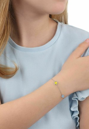 Giovane persona che indossa una maglietta a maniche corte a coste di colore azzurro chiaro e un braccialetto d'argento con ciondoli a forma di sole giallo e fiore blu sul polso sinistro.