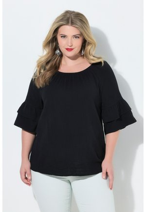 Blonde vrouw met los haar, zilveren oorbellen, een zwarte top met ruches aan de mouwen en lichtgekleurde broek tegen een neutrale achtergrond.