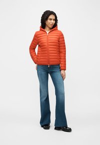 Γυναίκα που στέκεται με τα χέρια στις τσέπες, φοράει πορτοκαλί καπιτονέ μπουφάν με κουκούλα, μπλε ξεχειλωμένο τζιν και μαύρα παπούτσια σε λευκό φόντο.