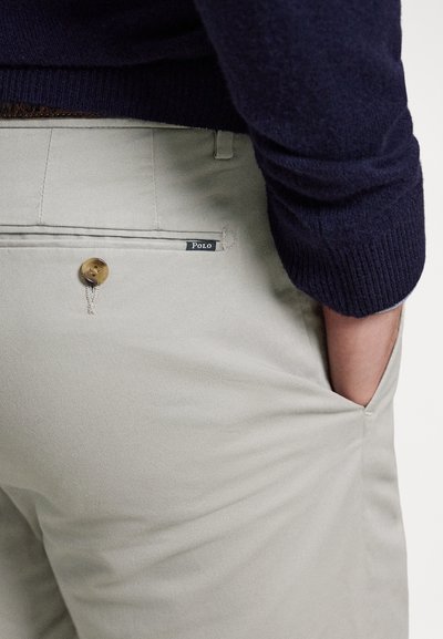 Beige byxor av bomullsblandningstyg, med bakficka med en bronzefärgad knapp och en synlig "Polo"-etikett, kombinerade med en marinblå tröja.