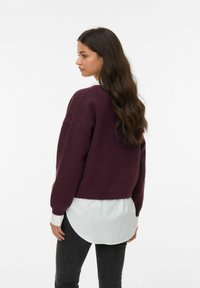 Pull en maille bordeaux à manches longues, à coupe décontractée, avec des poignets côtelés, superposé sur une chemise blanche à col. Tissu texturé.