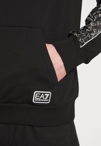 Svart hoodie med en framficka och EA7-logotyp. Har mönstrade ärmar och ribbade mudd. Materialet verkar slätt och stretchigt.