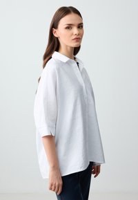 Camicia bianca con bottoni, realizzata in un tessuto testurizzato, con colletto e maniche a tre quarti. Presenta delle fessure laterali e una vestibilità rilassata per il massimo comfort.