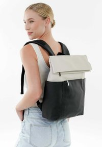 Mochila con fondo negro, tapeta superior beige y accesorios de metal plateado. Está hecha de material texturizado, con correas ajustables negras y un bolsillo con cremallera.