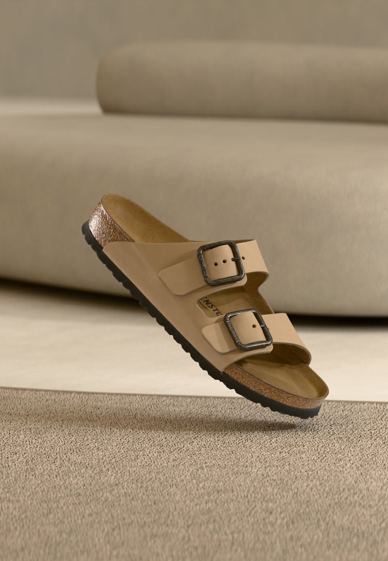 Ταμπά σανδάλι slide με δύο ρυθμιζόμενες αγκράφες και πάτο από φελλό, αιωρείται πάνω από μια υφασμάτινη μπεζ μοκέτα.