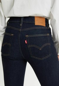 Jean en denim bleu foncé à taille haute, avec des coutures jaunes, deux poches arrière et un patch de marque en cuir beige à la taille.