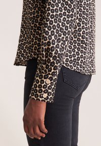 Blusa nera e crema con motivo floreale, maniche lunghe e polsini con bottoni, abbinata a jeans scuri. Tessuto dalla consistenza liscia.