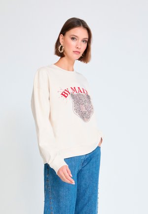 Mujer con cabello corto castaño, que lleva grandes pendientes dorados, sudadera crema con texto rojo y gráfico de tigre, y vaqueros azules con tachuelas laterales.