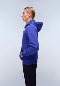 Felpa blu royal realizzata in tessuto morbido, con tasca a marsupio, polsini a costine e cappuccio con coulisse. Indossata con pantaloni neri.