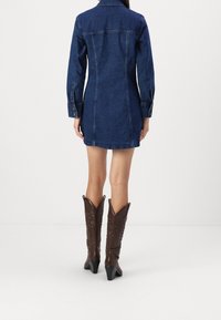 Vestido de denim con mangas largas y un cuello puntiagudo. Presenta una silueta ajustada y un ligero detalle de costura. Se lleva con botas de cowboy marrones.