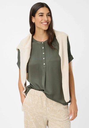 Olivgrüne kurzärmelige Bluse mit Knopfdetails, kombiniert mit hellbeigen gemusterten Hosen und einem kremfarbenen Strickpullover, der über die Schultern drapiert ist.