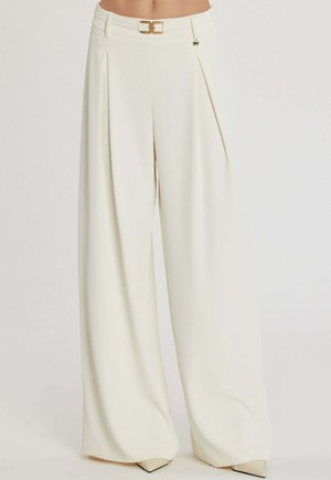 Pantaloni bianchi a gamba larga con pieghe, vita alta, dettaglio cintura con fibbia dorata, indossati con scarpe beige a punta.