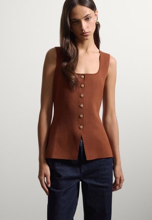 Mujer con top marrón sin mangas con botones delante y pantalones azul oscuro, de pie frente a un fondo claro y liso.