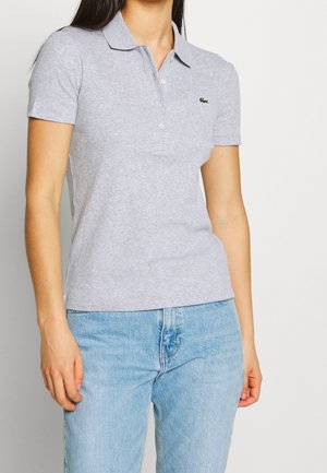 Vrouw in een lichtgrijze polo met korte mouwen, knopen en logo op de borst, gecombineerd met lichtblauwe denim jeans.