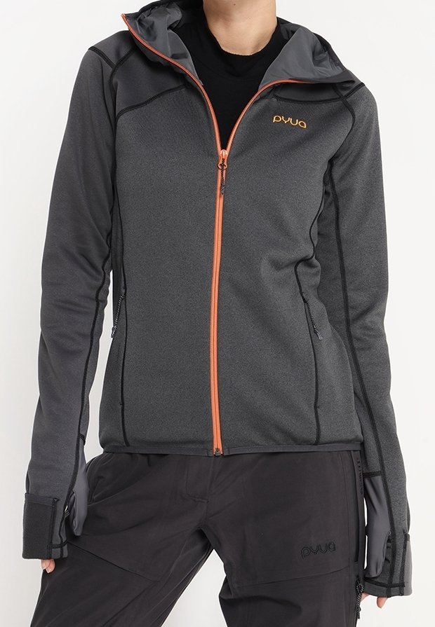 Personne portant une veste grise ajustée à capuche avec une fermeture éclair orange et un logo, accompagnée d'un pantalon noir et de gants, debout avec les bras détendus.