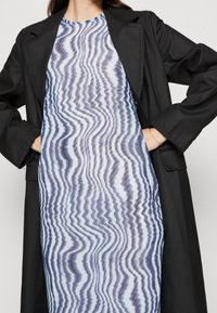 Veste noire sur une robe bleu clair présentant un motif ondulé texturé bleu et blanc. La robe a un col rond et une coupe ample.