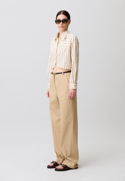 Maglietta a righe beige e bianca, tagliata, pantaloni beige a vita alta, sandali marroni con fibbie. La modella è in piedi davanti a uno sfondo semplice.