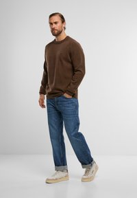 Brauner Strickpullover, blaue Jeans, weiße und beige Sneakers. Lockere Passform mit rundem Hals und gerippten Bündchen. Stehend vor einem schlichten Hintergrund.
