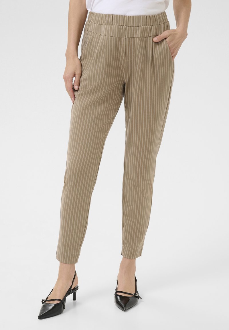 Pantalons rayés beiges avec une taille élastique, dotés de poches latérales et d'une coupe fuselée. Portés avec des chaussures à talons noires.