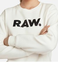 Osoba se založenýma rukama, oblečená v krémovém svetru s tučným černým nápisem "RAW." přes hruď.