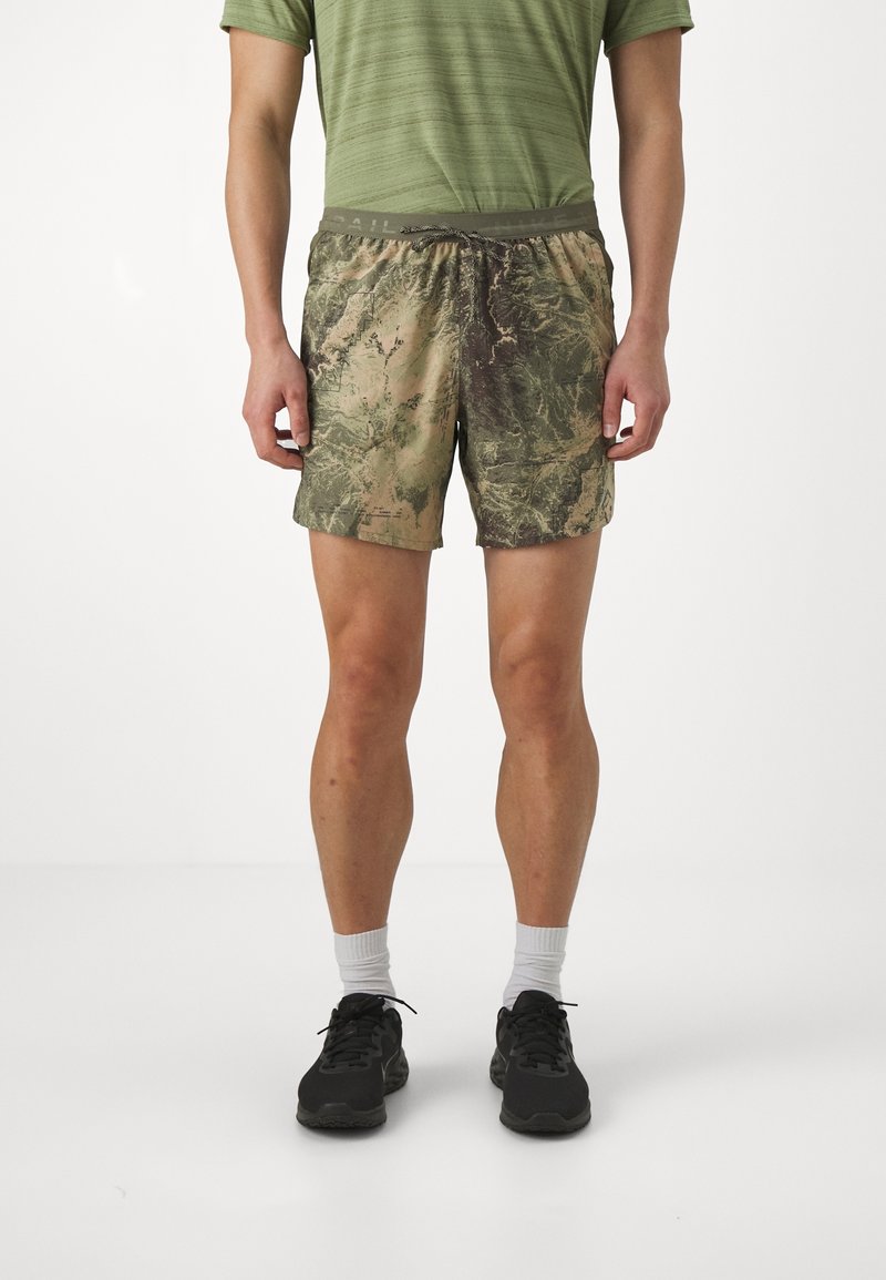 Homme portant un short vert à motif camouflage, un t-shirt vert olive, des chaussettes blanches et des baskets noires, debout devant un fond blanc uni.