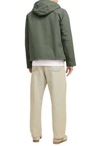 Giacca impermeabile verde con cappuccio, indossata sopra una camicia bianca, abbinata a pantaloni beige chiaro e scarpe color cuoio. La vista posteriore mette in evidenza la semplicità.