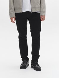 Svarta figurnära byxor kombinerade med en ljusgrå zip-up jacka över en vit t-shirt. Skorna är svarta läderskor med en texturerad sula.