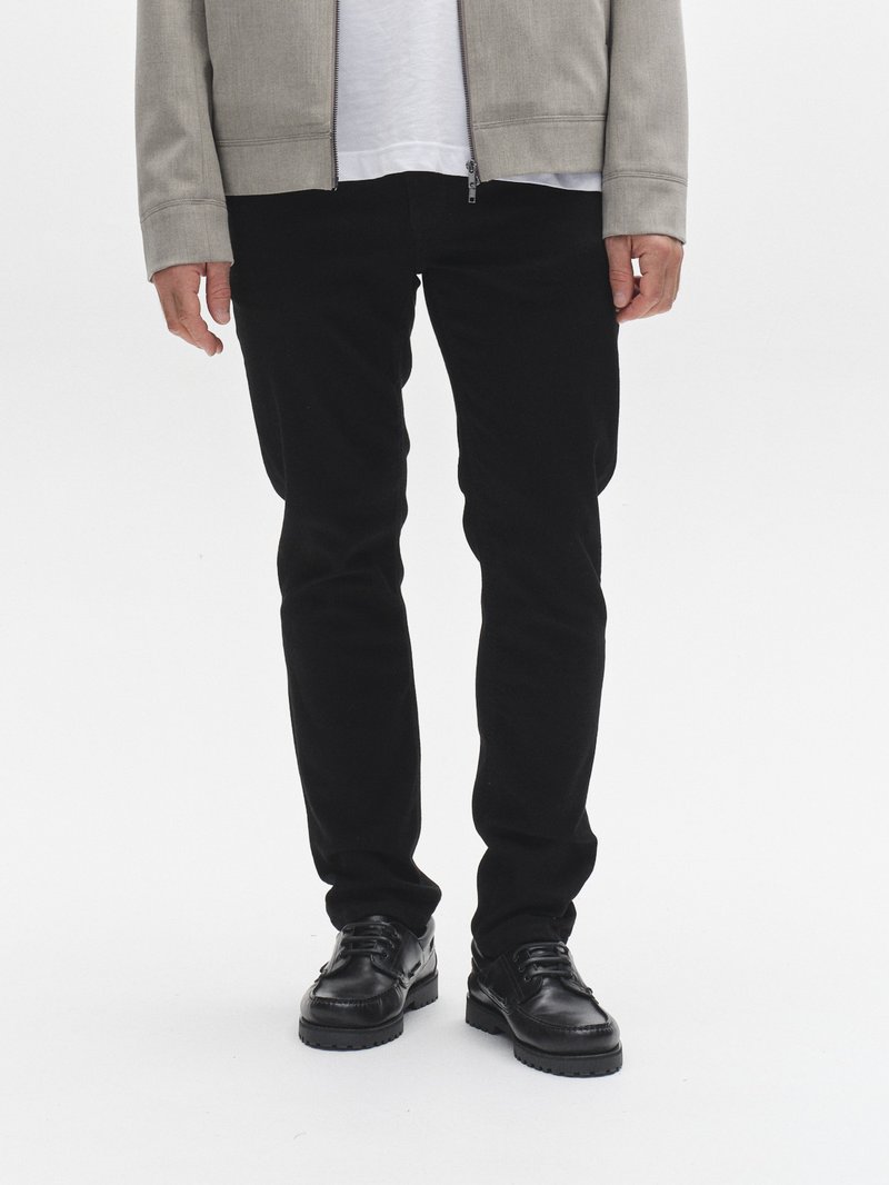 Svarta figurnära byxor kombinerade med en ljusgrå zip-up jacka över en vit t-shirt. Skorna är svarta läderskor med en texturerad sula.