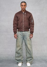 Bomber jacket in suede marrone con zip frontale, polsini a coste e dettagli con toppe; abbinato a jeans a gamba larga blu sbiaditi e sneakers bianche.