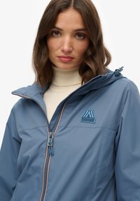 Niebieska kurtka zapinana na zamek, wykonana z lekkiego materiału, z kapturem, haftowanym logo i kontrastowymi szwami wzdłuż zamka.