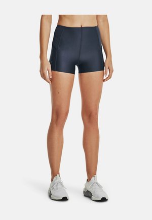 Kobieta w czarnych sportowych szortach i białych butach do biegania, stojąca na tle jednolitego białego tła.