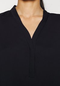 Blouse noire avec un décolleté en V, présentant des détails subtils de couture le long de l'encolure. Texture lisse, tissu léger.