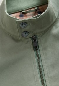 Groene stoffen jas met een hoge kraag, voorzien van twee knopen en een rits aan de voorkant. Van binnen heeft de jas een geruit patroon in verschillende bruintinten.
