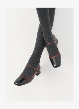 Chaussures Mary Jane en cuir bordeaux avec un contrefort en verni noir et une fine sangle à la cheville ; portées avec des chaussettes côtelées gris foncé. Talon carré bas.