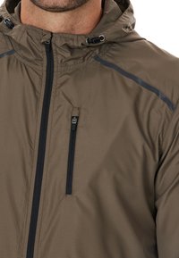 Braune wasserdichte Jacke mit einer Reißverschlusstasche auf der Brust, kontrastierendem schwarzen Reißverschluss und verstellbare Kapuze. Verfügt über strukturiertes Gewebe und ein dezentes Muster.