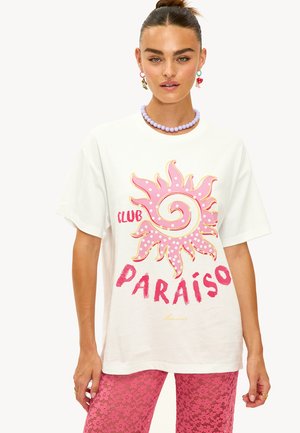 Camiseta blanca de gran tamaño con un gráfico de sol rosa, texto "CLUB PARAÍSO" en un vibrante color rosa y acentos dorados. La modelo lleva un collar de cuentas en color lavanda.