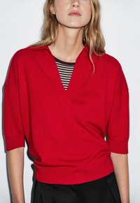 Czerwony sweter z dzianiny, z dekoltem w kształcie litery V i krótkimi rękawami, nałożony na czarno-biały, paskowany T-shirt, charakteryzujący się luźnym krojem i prążkowanym dołem.