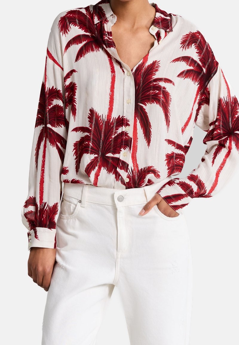 Camicia a maniche lunghe con bottoni, realizzata in tessuto bianco con stampe di palme rosse. Il colletto è aperto e viene indossata con pantaloni bianchi.