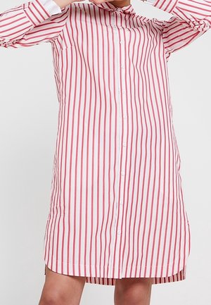 Personne portant une robe à manches longues boutonnée avec des rayures verticales rouges et blanches, tenant un cintre près du col.