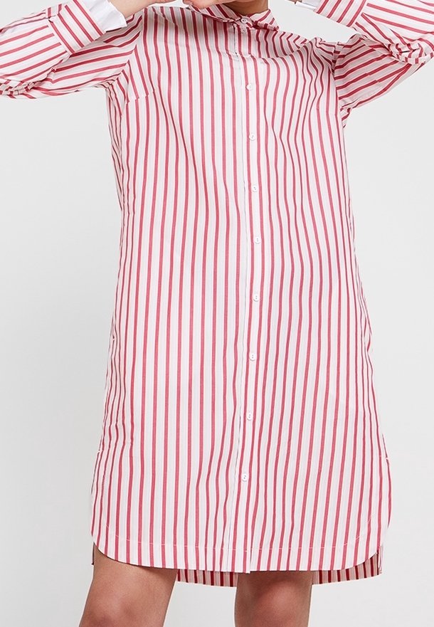 Personne portant une robe à manches longues boutonnée avec des rayures verticales rouges et blanches, tenant un cintre près du col.