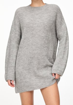 Person iført en lang, ribstrikket grå sweaterkjole med lange ærmer, stående mod en simpel hvid baggrund.