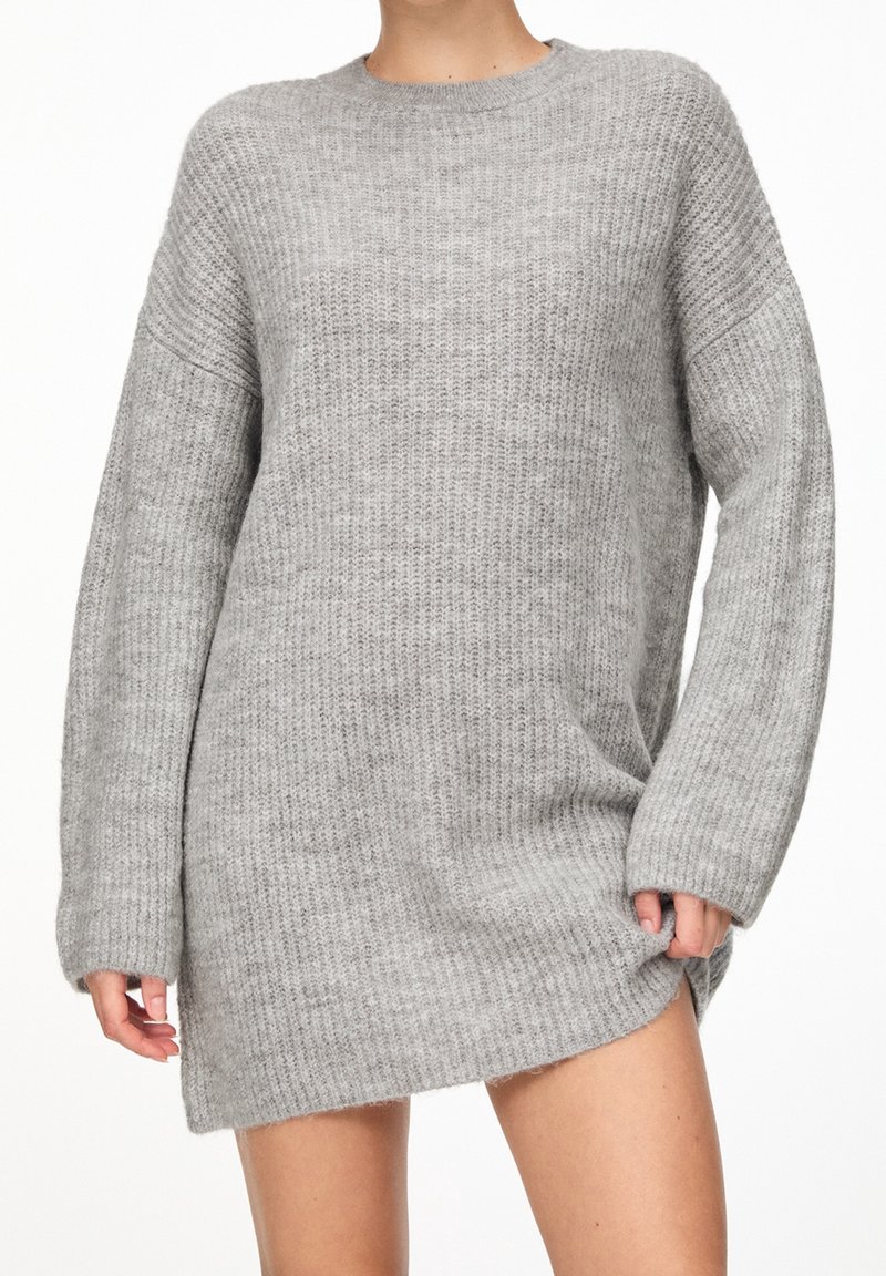 Personne portant une robe-pull grise longue en maille côtelée à manches longues, debout devant un fond blanc uni.