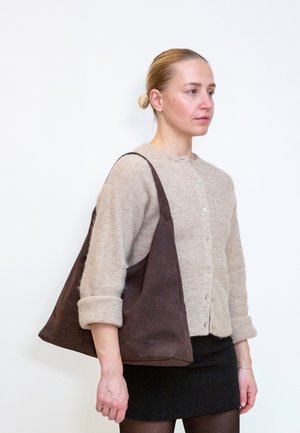 Frau mit blondem Haar zu einem Dutt gebunden, trägt eine beige Strickjacke und einen schwarzen Rock und trägt eine große dunkelbraune Umhängetasche.