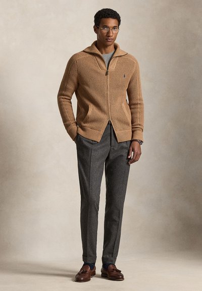 Bărbat cu ochelari, purtând un pulover tricotat camel cu fermoar, pantaloni gri și mocasini maro din piele, stând cu mâinile în buzunare.